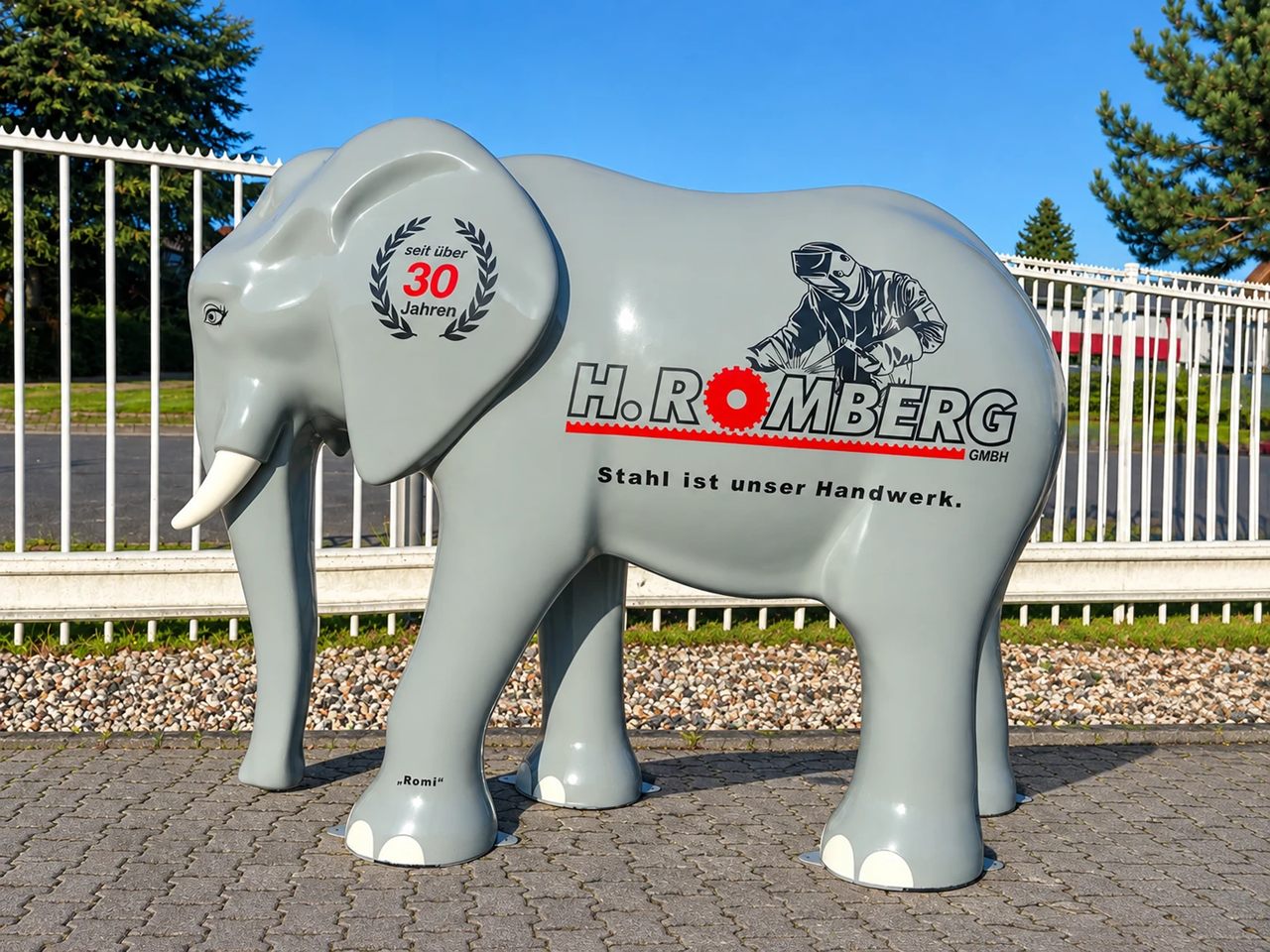 Elefantenfigur Romi auf einem gepflasterten Platz vor einem weißen Zaun, bedruckt mit dem Logo der H. Romberg GmbH, einem Schweißer, dem Siegel „seit über 30 Jahren“ und dem Satz „Stahl ist unser Handwerk.“